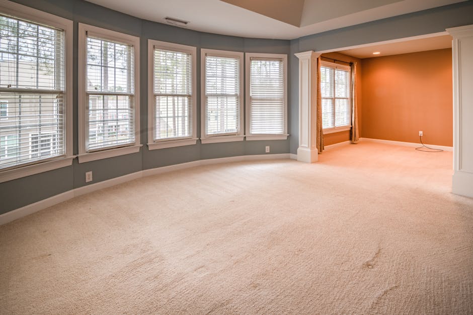 A man with grey hair and a beard is performing surface cleaning in a bright, well-lit living room. He is using a vacuum cleaner on a yellow and white patterned rug that lies on polished dark wooden fl