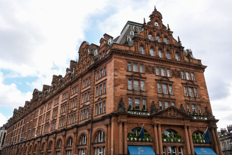 Exterior view of a historic Victorian-style building constructed from red sandstone, featuring ornate architectural details, multiple arched windows, and decorative stone carvings. The building's uppe